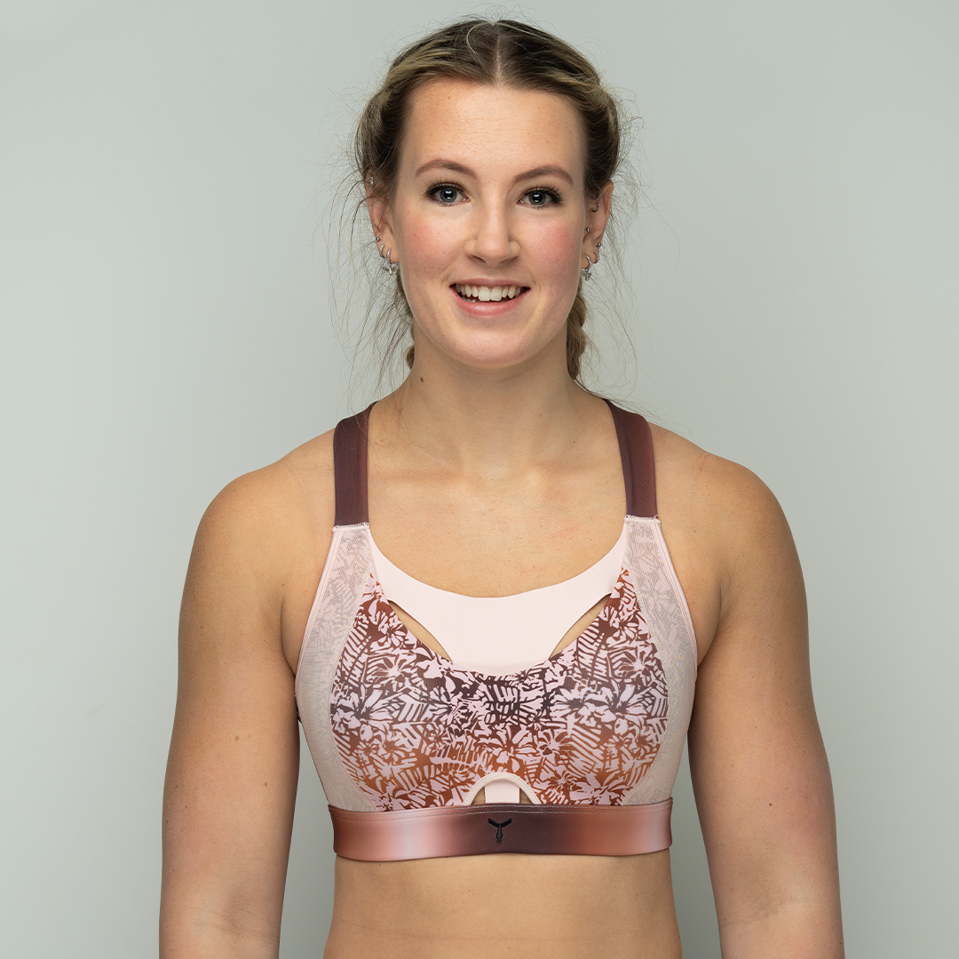 Model wearing the Tigers Eye Orchid sports bra, standing against a sleek, minimal grey background, smiling at the camera. Designed for ultimate support, breathability, and comfort during intense workouts.