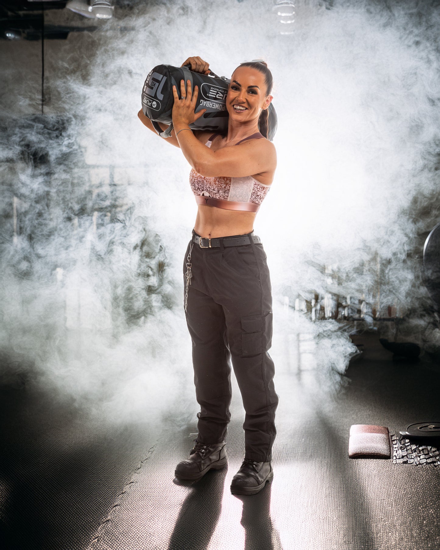 Female prison office wearing a pink Tigers Eye Ultimate Bra lifting a sand bag on her shoulder in a dark gym setting with smoke effects.